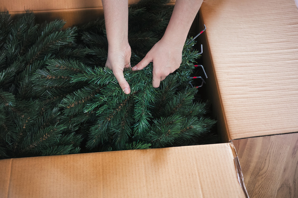 Žena rukami vyťahuje z veľkej kartónovej škatule konáre umelého vianočného stromčeka